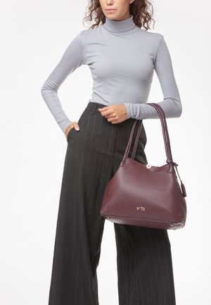 Donna con dolcevita grigio e pantaloni neri a righe sottili che tiene una grande borsa a mano in pelle bordeaux con logo dorato.