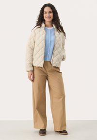 Prešívaná krémová bunda so zipsom vpredu, v kombinácii s svetlomodrým kardigánom a béžovými nohavicami s širokým strihom. Hnedé topánky dokončujú outfit.