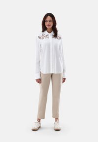 Camicia bianca con bottoni, con dettagli in pizzo sul colletto, abbinata a pantaloni beige e sneakers di colore chiaro. Texture del tessuto liscia.