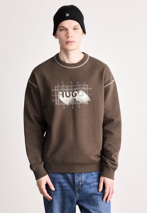Brauner Sweatshirt mit weißem Grafikdesign "HUGO", vor einem Gittermuster platziert. Gerippte Bündchen und runder Halsabschluss, kombiniert mit blauen Jeans.