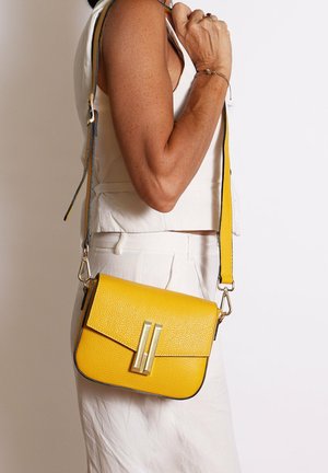 Donna che indossa un abito bianco con una borsa a tracolla gialla dalla texture, caratterizzata da una chiusura dorata, portata sulla spalla contro uno sfondo semplice.