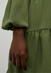 Vihreä teksturoitu mekko, jossa on kerätty vyötärö ja leveät hihat, sekä näkyvä käsi pitämässä kangasta. Lähikuva materiaalin yksityiskohdista.