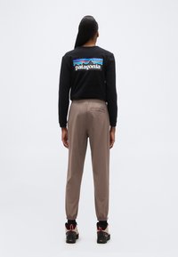 Άτομο που φοράει μαύρο φούτερ Patagonia και μπεζ παντελόνι, όρθιο με την πλάτη στραμμένη σε απλό φωτεινό φόντο.