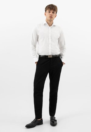 Young man standing with hands in pockets, wearing a white button-up shirt, black pants, black belt, and black dress shoes against a white background.