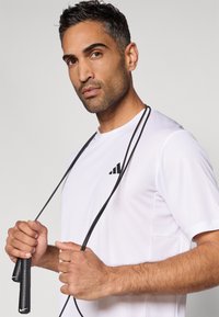 Camiseta desportiva branca com acabamento texturizado e logótipo preto; indivíduo segura uma corda de saltar preta com pegadores texturizados, exibindo uma expressão concentrada.