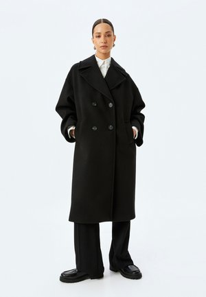 Manteau en laine noir à double boutonnage avec grands revers, deux poches avant et tissu texturé. Assorti à un pantalon noir à jambes larges et des chaussures à plateforme.