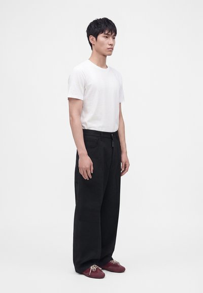Homme debout portant une chemise blanche à manches courtes, un pantalon large noir et des chaussures rouges avec des lacets blancs sur un fond blanc uni.