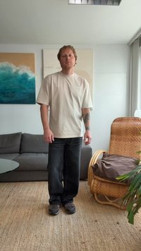 T-shirt beige à manches courtes avec une coupe décontractée, associé à un jean en denim foncé et des chaussures de sport noires. Dans un intérieur moderne avec un canapé.