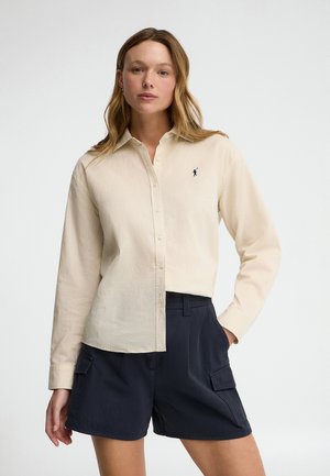 Frau trägt ein cremefarbenes Hemd mit Knöpfen und kleinem besticktem Logo sowie marineblaue Cargo-Shorts, steht mit einer Hand in der Tasche.