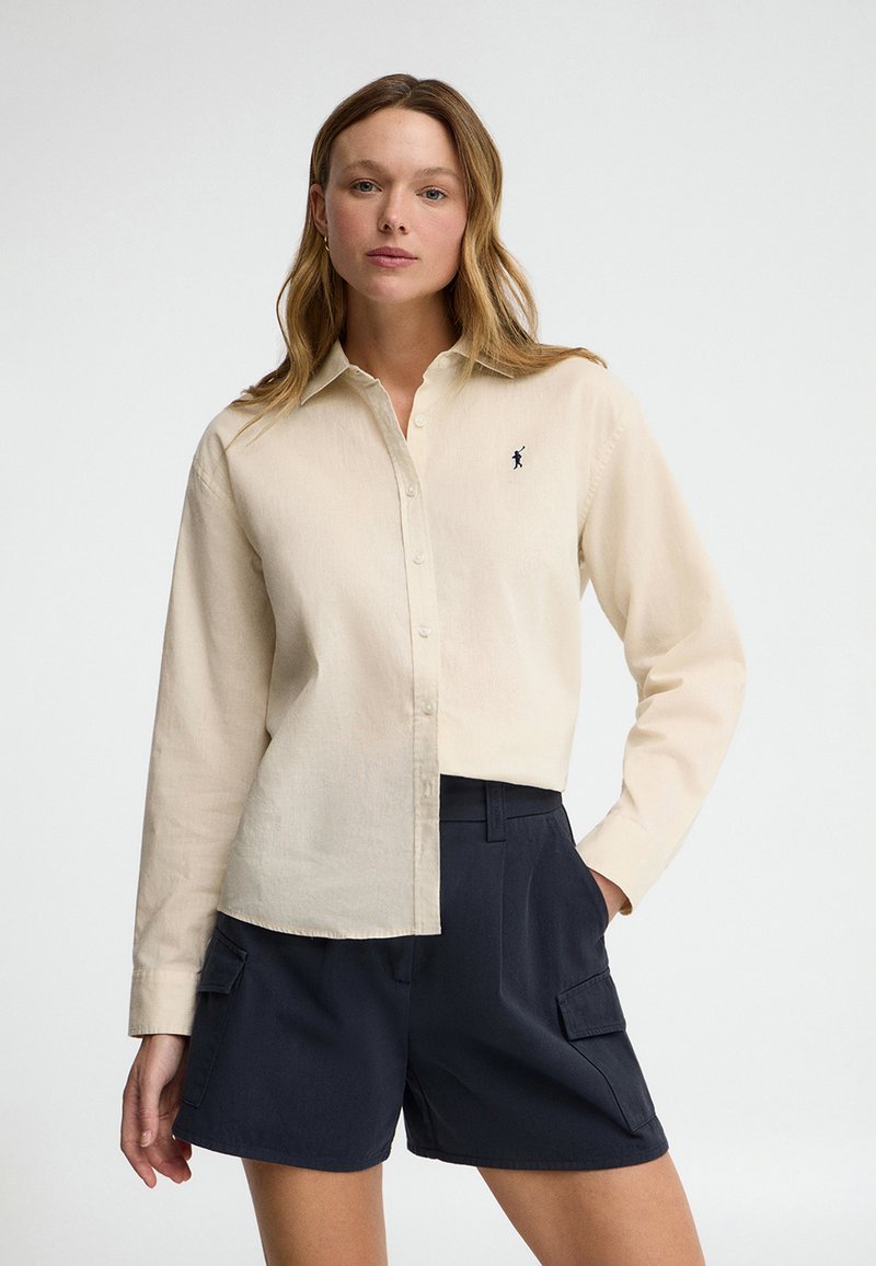 Vrouw draagt een crème knopenblouse met een klein geborduurd logo en marineblauwe cargoshorts, staand met één hand in de zak.