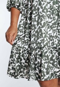 Robe florale verte et blanche avec un ourlet en volants, présentant un tissu texturé et des manches bouffantes. Le design inclut des motifs de feuilles distincts.