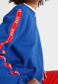 Blå sweatshirt med röda "NIKE"-logotypdetaljer längs ärmarna, med rund hals samt strukturerad tyg.