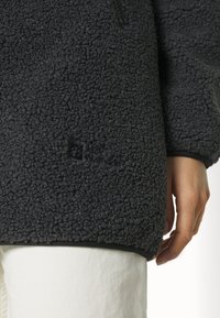 Dunkelgraue Fleecejacke mit strukturierter Oberfläche, ausgestattet mit einem Logo unten rechts und schwarzem Saum sowie Bündchen.
