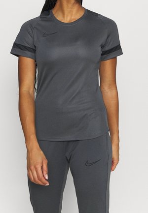 Femme portant un t-shirt et un pantalon de sport Nike gris foncé, debout devant un fond clair uni, avec un bras détendu sur le côté.