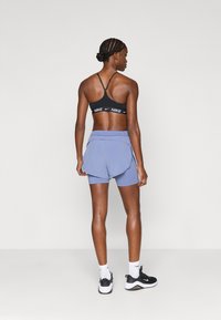 Μαύρο αθλητικό σουτιέν με λευκό λογότυπο Nike, λιλά σορτς με επιμελημένο σχέδιο και μαύρα αθλητικά παπούτσια με λευκές λεπτομέρειες, απεικονιζόμενα από πίσω.