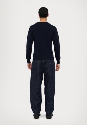 Homme portant un pull noir et un jean bleu foncé ample, debout de dos face à un fond clair uni.