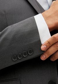 Veste de costume grise en tissu texturé, avec un doublure de manchette blanche et quatre boutons noirs sur la manche pour la fermeture.