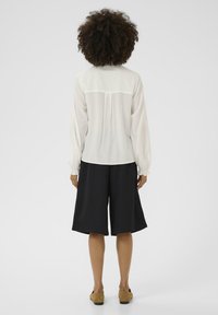 Blusa bianca a maniche lunghe con polsini elastici, abbinata a pantaloni corti neri fino al ginocchio. Tessuto dalla consistenza liscia e vestibilità comoda visibile dal retro.