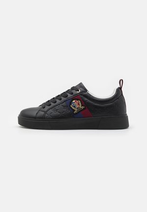 Black leather sneaker featuring a textured surface, lace-up design, rubber sole, and side stripe with an embroidered crest in red and blue.