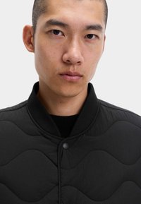 Svart vadderad jacka med ribbat krage, med tryckknappsstängning och vågiga texturmönster på tyget.