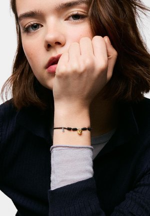 Jeune femme aux cheveux bruns mi-longs, reposant son menton sur son poing, portant un bracelet noir à perles avec un petit pendentif cœur en or.