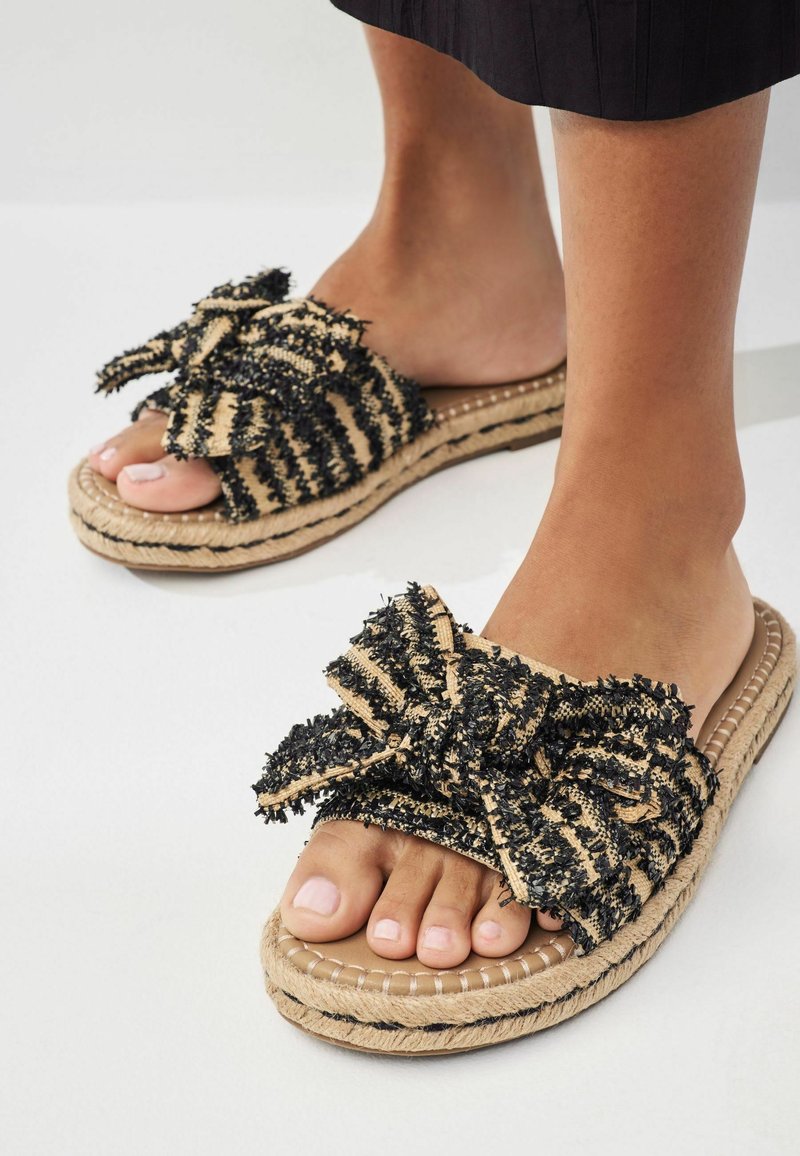 Pieds portant des sandales à enfiler tissées beige et noir avec des détails de nœud épais texturés et une semelle en jute tressée sur une surface blanche.