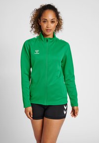 Grøn atletisk jakke med en frontlynlås, høj krave og logo. Har lange ærmer og sorte shorts med hvide chevron-detaljer.