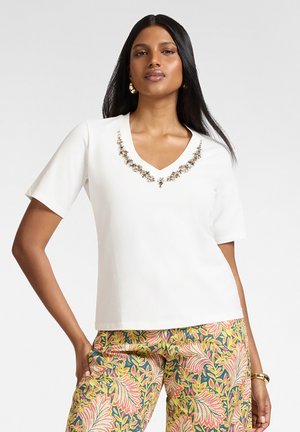 Vrouw draagt een wit V-hals shirt met een met kralen versierde halslijn en kleurrijke bloemenbroek, staand met één hand op de heup.