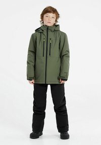Veste imperméable verte avec fermeture éclair à l'avant et deux poches, associée à un pantalon noir et des bottes de neige, dotée d'une capuche et de poignets réglables.