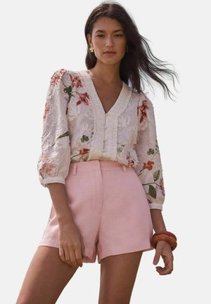 Woman with long dark hair wearing floral lace blouse with puff sleeves and light pink high-waist shorts, standing with one hand relaxed.