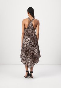 Femme aux cheveux tressés porte une robe sans manches, dos croisé, imprimé léopard et des sandales noires à talons hauts, debout sur un fond uni.