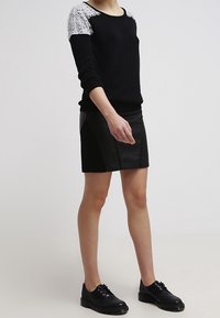 Personne portant un pull noir avec des épaules en dentelle blanche, une mini-jupe en cuir noir et des chaussures noires à lacets, debout contre un fond uni.