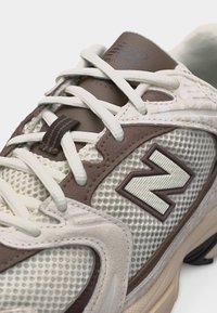 Prim-plan cu un pantof sport bej și maro din plasă, cu șireturi albe și logo „N” texturat pe lateral.