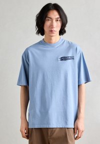 Tricou supradimensionat de culoare albastru deschis, fabricat din bumbac, cu mâneci scurte și un grafic imprimat în zona stângă a pieptului. Textură netedă.