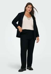 Černý oblekový blazer přes smetanový top s krajkovým lemem, spárovaný s černými kalhotami a lesklými černými botami. Jednoduchý, strukturovaný design.