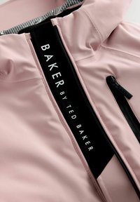 Gaiši rozā jaka ar augstu apkakli, melnu rāvējslēdzēju un zīmola etiķeti. Raksturīga gluda tekstūra un kontrastējoši melni akcenti.