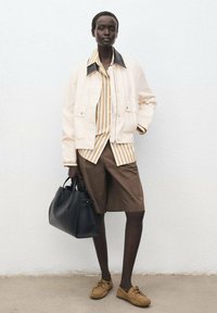 Light beige jacket with dark collar, striped shirt, brown shorts, tan shoes, and a black handbag. Smooth textures, casual fit.