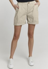 Shorts en coton beige avec des manches repliées, des poches avant et une fermeture à bouton. Portés avec des baskets blanches, se tenant sur un fond uni.