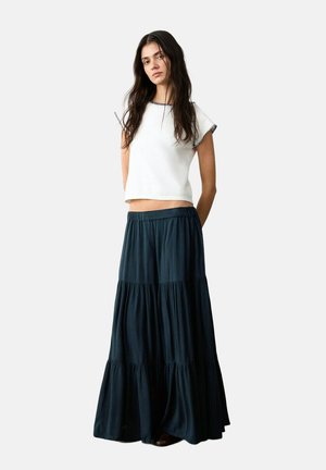 Giovane donna con capelli lunghi e scuri che indossa un top corto bianco a maniche corte e una lunga gonna maxi fluente e a balze scura, su sfondo neutro.