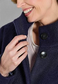 Marineblauer Wollmantel mit hohem Kragen, versehen mit schwarzen Knöpfen und einer strukturierten Oberfläche. Eine Hand hält sanft das Kragenelement.