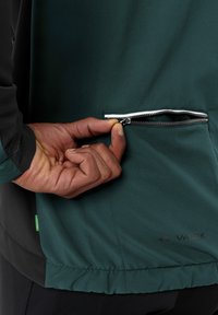 Giacca verde scuro con base nera, dotata di tasca con zip e orlo con cordino. Tessuto testurizzato con un aspetto liscio.