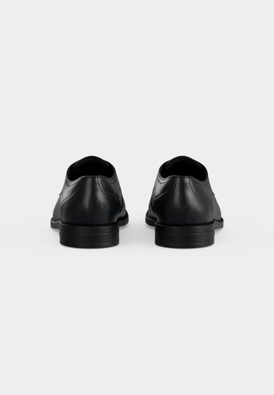 Paire de chaussures habillées en cuir noir vues de dos, sur un fond gris clair uni avec des talons bas.
