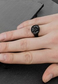 Anillo negro hecho de material mate con un diseño en relieve, reposando sobre una mano de piel clara, sobre una superficie texturizada.