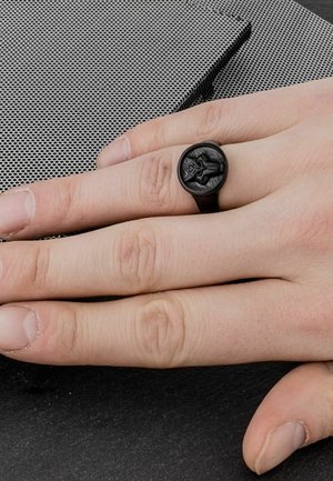 Anello nero realizzato in materiale opaco con un design in rilievo, poggiante su una mano dalla pelle chiara, su una superficie texture.