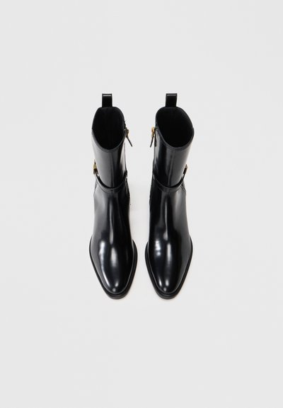 Par de brillantes botas de tobillo negras con puntera afilada y cremalleras, mostradas desde arriba sobre un fondo blanco.