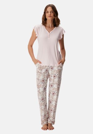 Vrouw staat blootsvoets, draagt een lichtroze top met korte mouwen en witte losse broek met bloemmotief, handen in de zakken.