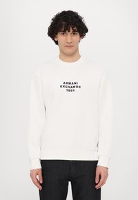 Hvid sweatshirt med ribbede manchetter og kant, der har sort broderet tekst "ARMANI EXCHANGE 1991" på forsiden. Glat tekstur.