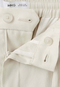 Gros plan sur la ceinture d'un pantalon beige montrant un bouton, une boutonnière, une fermeture éclair et une étiquette en tissu avec les détails de la marque et de la taille.