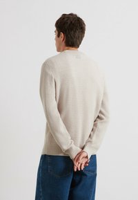 Beige strukturierter Pullover mit Rundhalsausschnitt und langen Ärmeln, kombiniert mit blauen Jeans. Der Pullover verfügt über ein dezentes Waffelmuster und gerippte Bündchen.