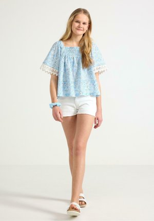 Teini-ikäinen tyttö kävelee sinikuvioinen pusero, jossa pitsihiha, valkoiset shortsit, valkoiset sandaalit ja sopiva sininen scrunchie ranteessa.